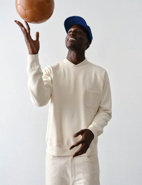A man wearing an undyed IOAN Pocket Sweatshirt and matching pants, with a blue cap, balancing a brown soccer ball on his outstretched hand against a plain light gray background.