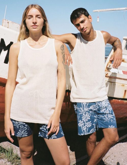 A man and a woman wearing matching off-white sleeveless tank tops and matching IOAN white floral boardshorts, standing outdoors in front of a boat.