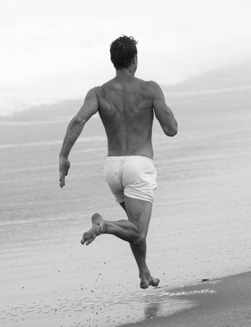 A black and white photograph of a man running barefoot on a beach, viewed from behind, wearing IOAN Jersey Boxers.