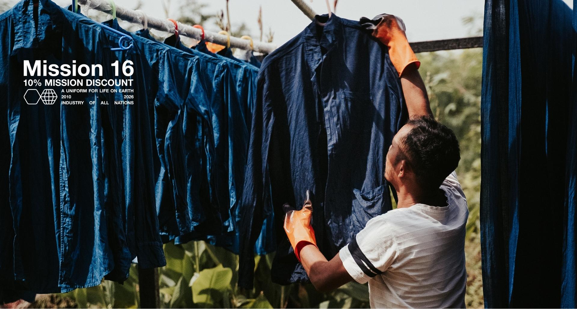 An IOAN dyemaster in India wearing orange gloves hangs a naturally dyed indigo shirt on an outdoor rack, surrounded by other indigo garments and green foliage.