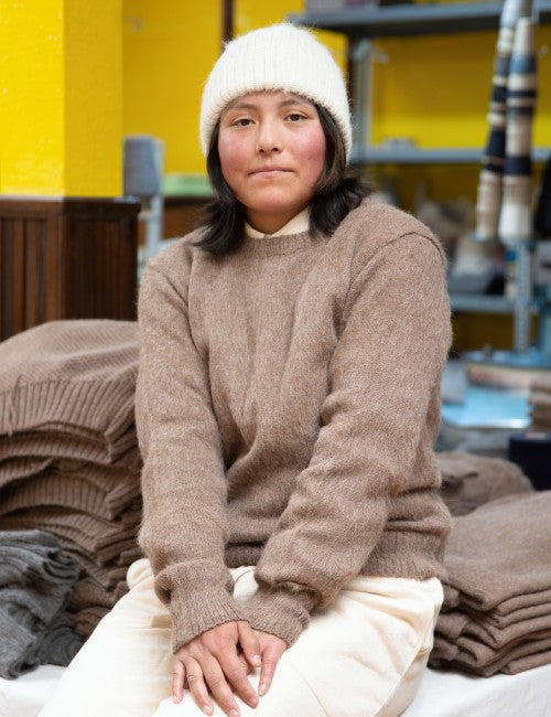 A person wearing a chunky knit taupe-brown sweater, cream beanie, and cream pants, sitting in a workshop with stacks of folded sweaters in the background.