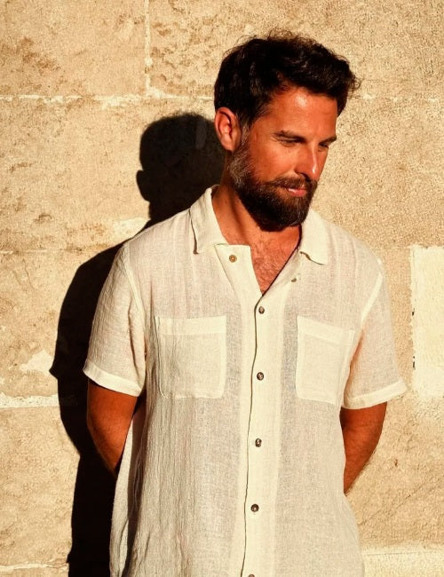 A man with a beard wearing a short-sleeve linen button-up shirt in a warm off-white color, standing against a textured stone wall with a strong shadow cast on it.