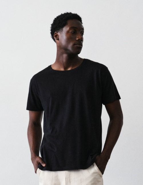 A man wearing a plain black short-sleeve crewneck t-shirt and off-white trousers, standing against a light gray studio background.