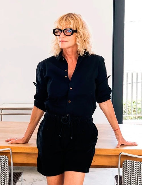 A woman with blonde curly hair and black glasses stands leaning on a wooden table, wearing a black button-up shirt with rolled sleeves and a matching black drawstring mini skirt.