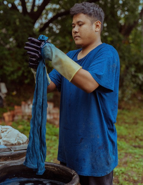 A man wearing a blue t-shirt and protective gloves holds up a dark blue fabric item over a basin outdoors.