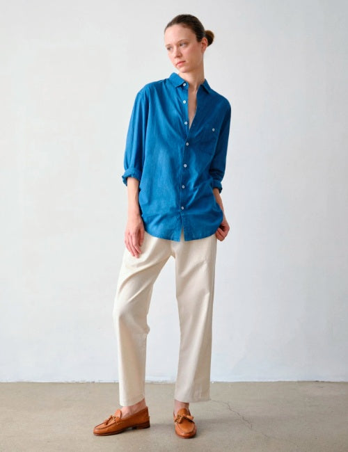 A model stands against a plain light gray wall wearing a bright blue button-up shirt, cream-colored trousers, and brown leather loafers.