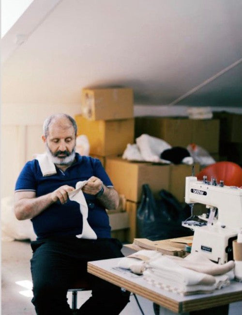 A bearded man in a blue shirt sits at a table in a workshop, handling white fabric pieces next to a white sewing machine. Cardboard boxes and bags are stacked in the background.