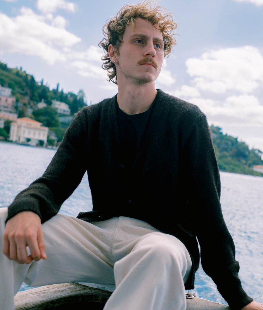 A young man wearing a black organic cotton sweater sitting on a wooden dock overlooking a body of water - Industry of All Nations