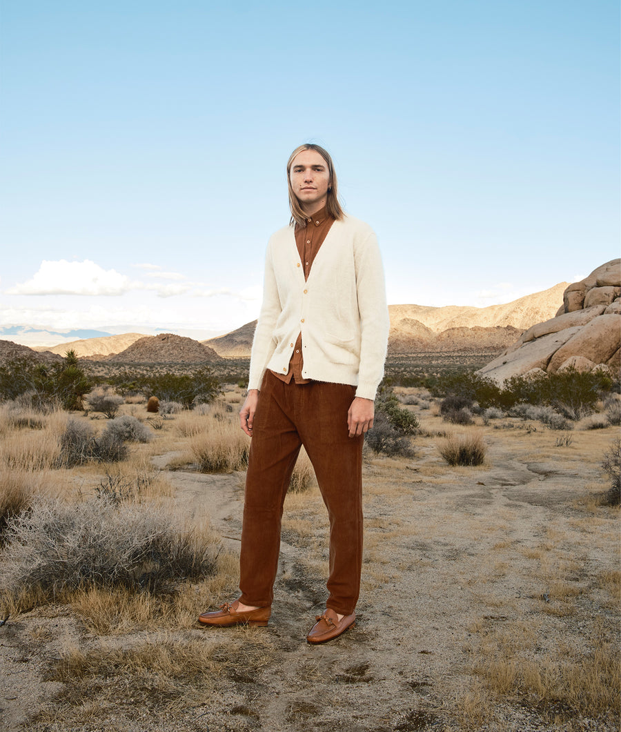 A person wearing a white cardigan, brown button-up shirt, and brown pants standing in a desert landscape - Industry of All Nations