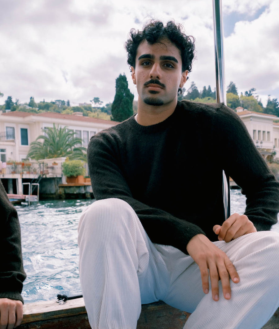 A man wearing a black sweater and white pants sitting on a wooden platform overlooking a body of water - Industry of All Nations