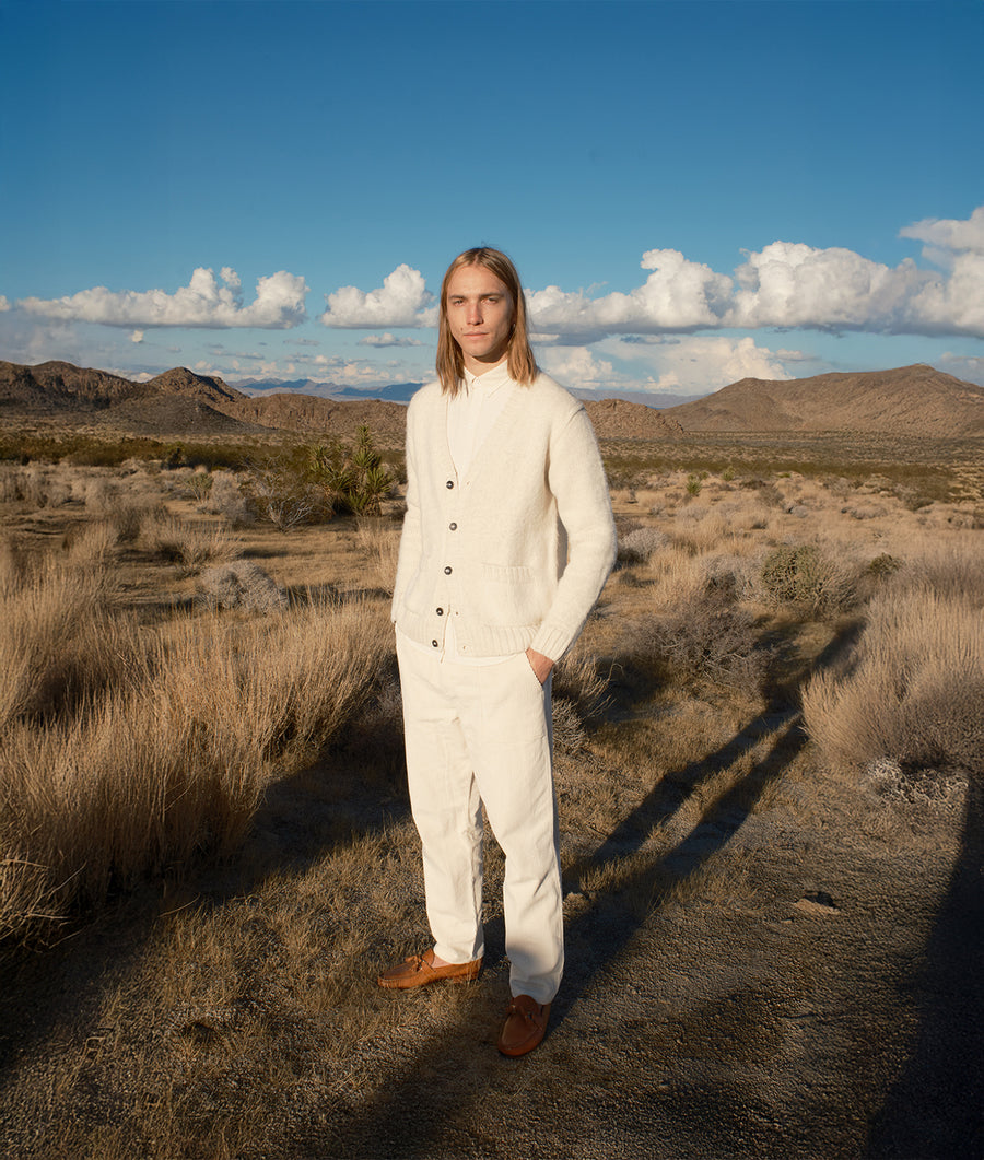 A person wearing a cream-colored sweater and pants, standing in a desert landscape - Industry of All Nations.