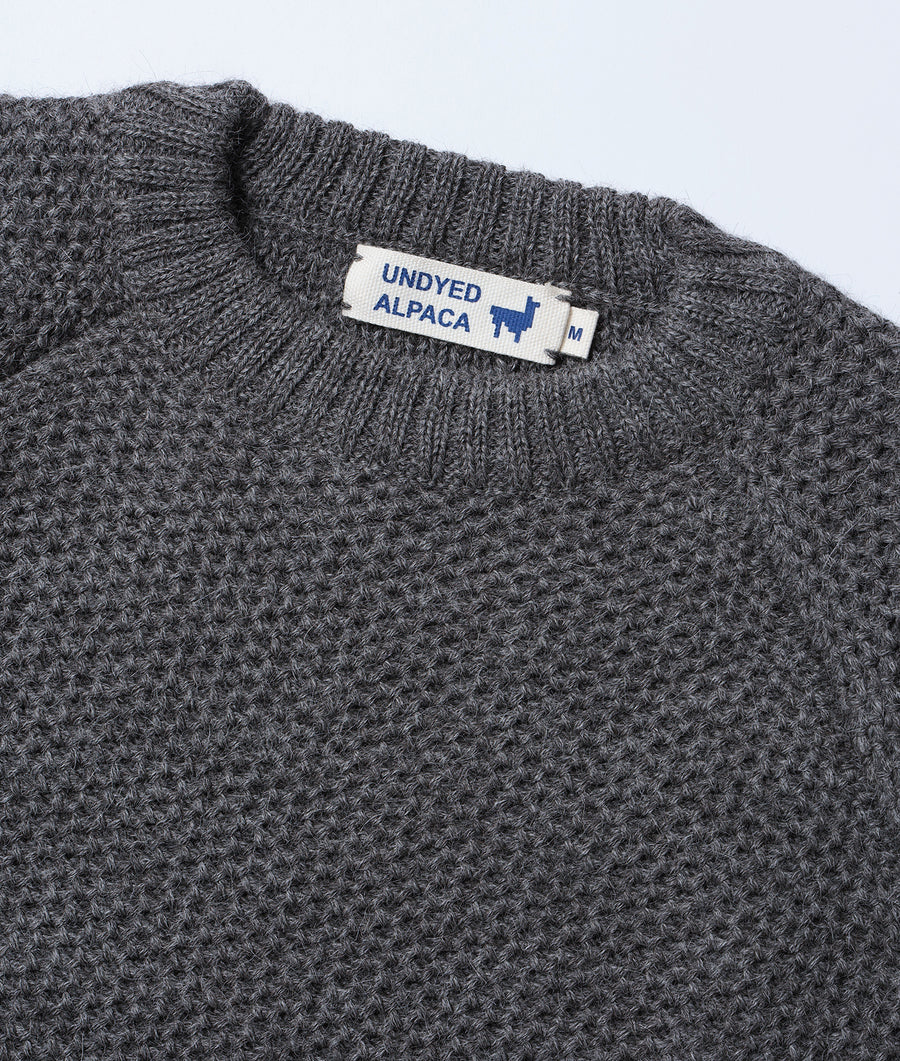 A close-up detail shot of a grey crocheted alpaca sweater with the Undyed Alpaca brand tag visible - Industry of All Nations