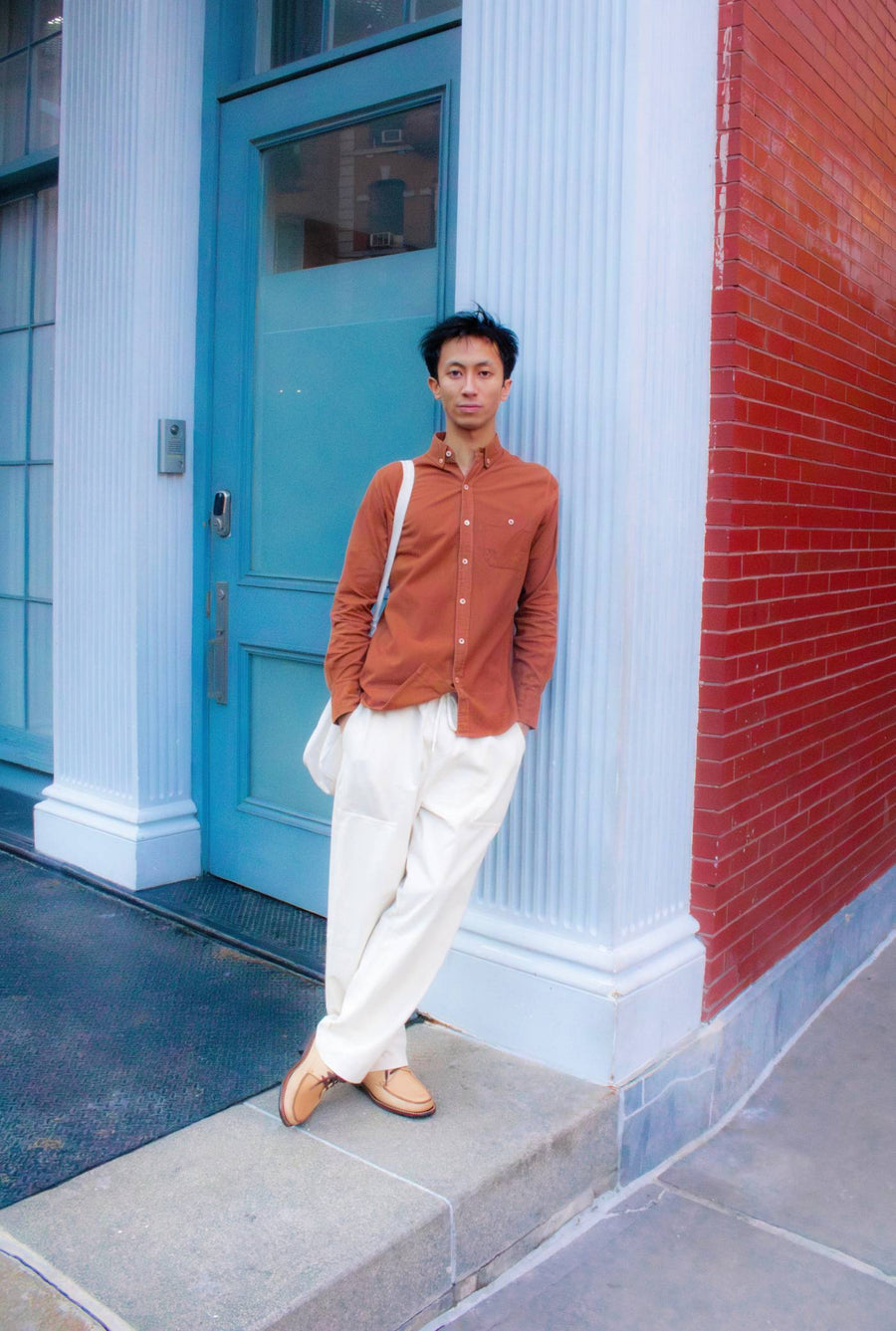 A person wearing an orange long-sleeved collared shirt and white pants, standing in front of a blue and red brick building - Industry of All Nations