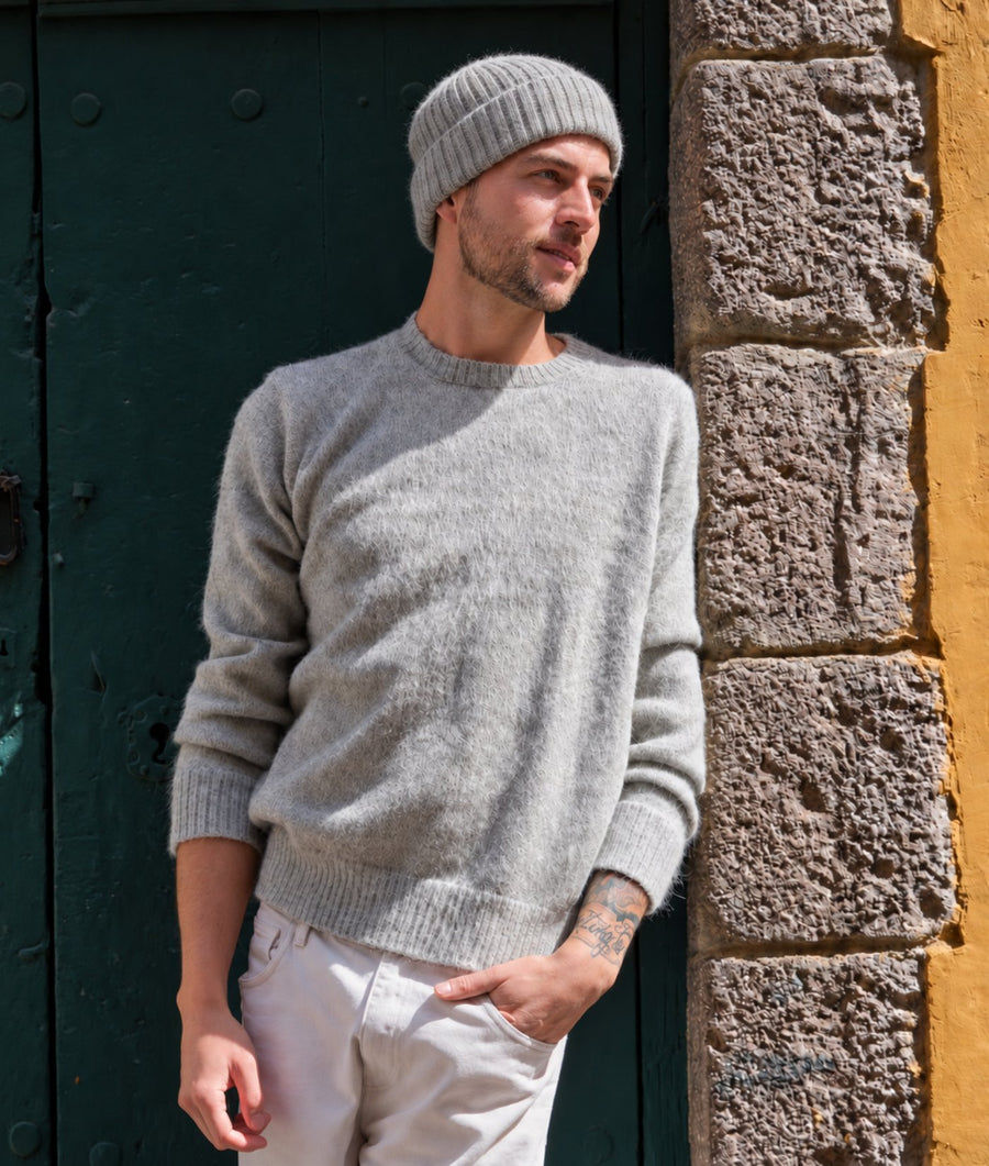 A man wearing a light gray knit crewneck sweater, a matching gray beanie, and off-white trousers, standing outdoors next to a dark green door and a textured stone wall.