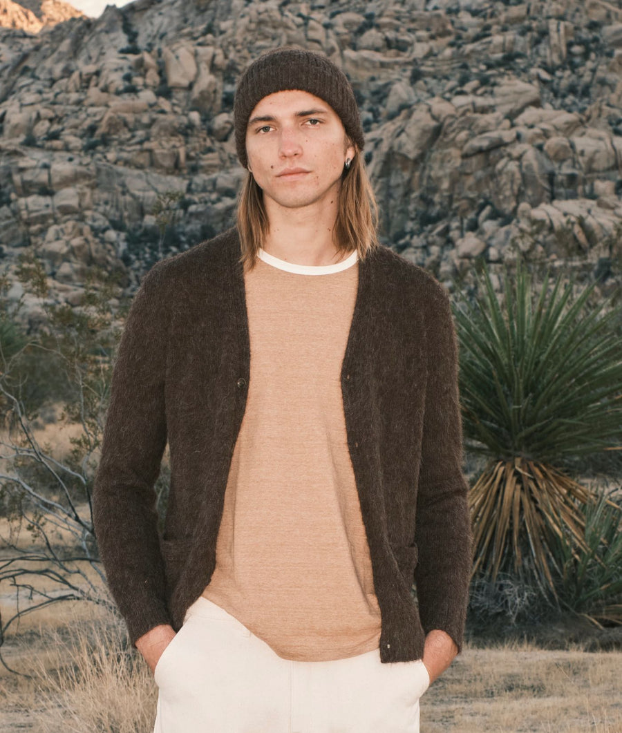 A man wearing a dark brown knit cardigan over a tan crewneck sweater, a dark brown beanie, and off-white pants, standing in a rocky desert landscape.