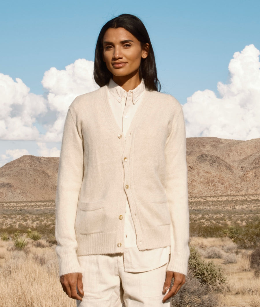 A woman stands outdoors in a desert landscape wearing a cream-colored knit cardigan over a matching collared shirt and light pants. The background features dry grass, rocky hills, and a blue sky with white clouds.