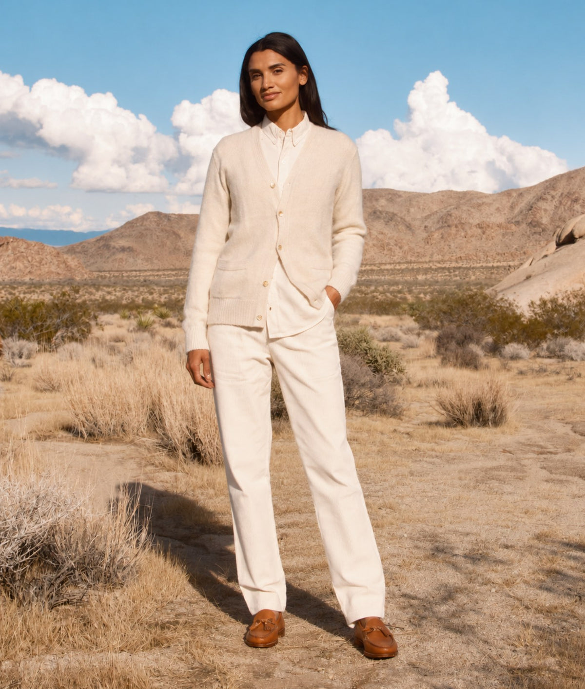 A woman stands in a desert landscape wearing a matching cream-colored knit cardigan and trousers set with brown suede loafers. The background features dry brush, distant mountains, and a blue sky with white clouds.