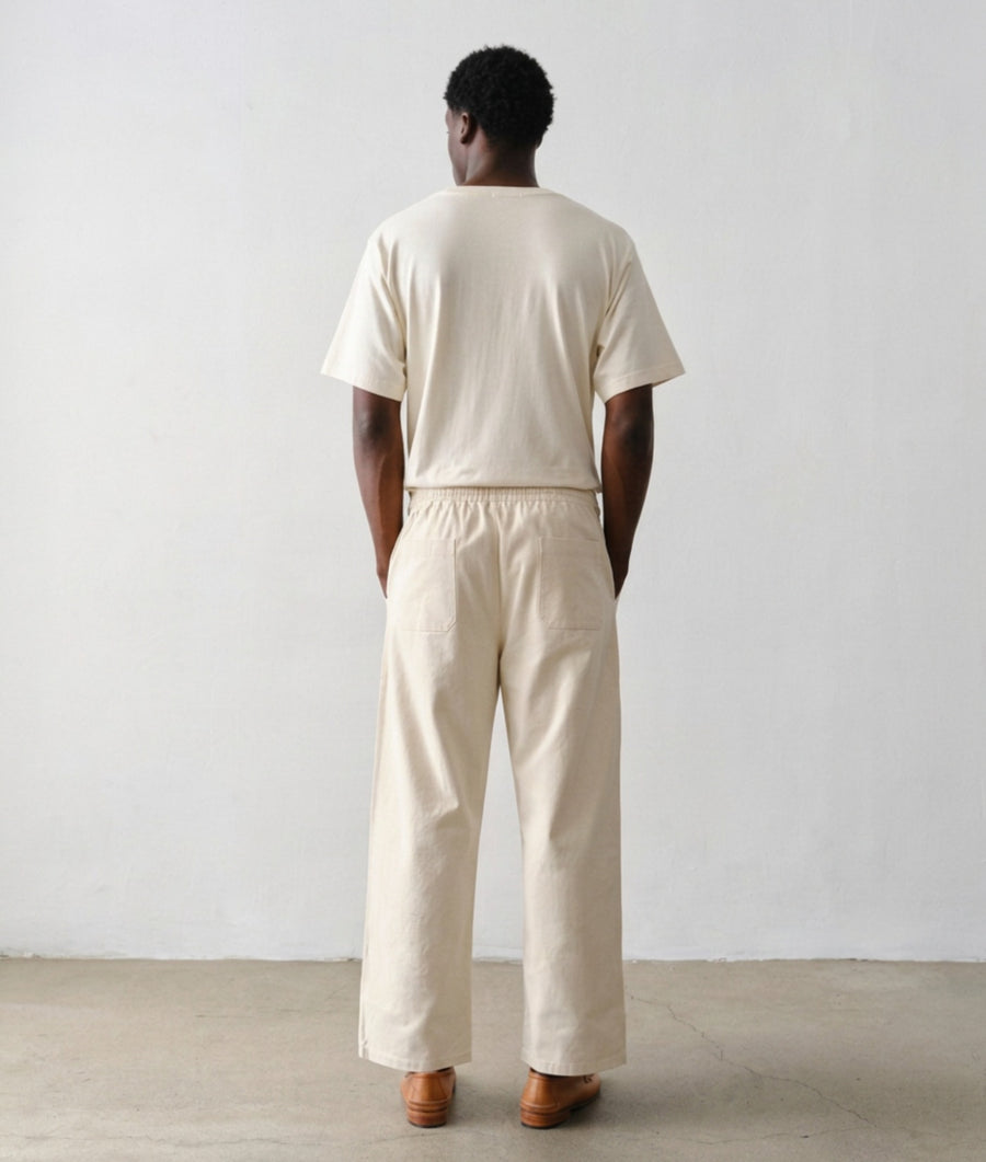 A man wearing an IOAN Clean Crewneck T-shirt with matching undyed Ango Pants, paired with brown leather loafers, standing against a plain light gray wall.