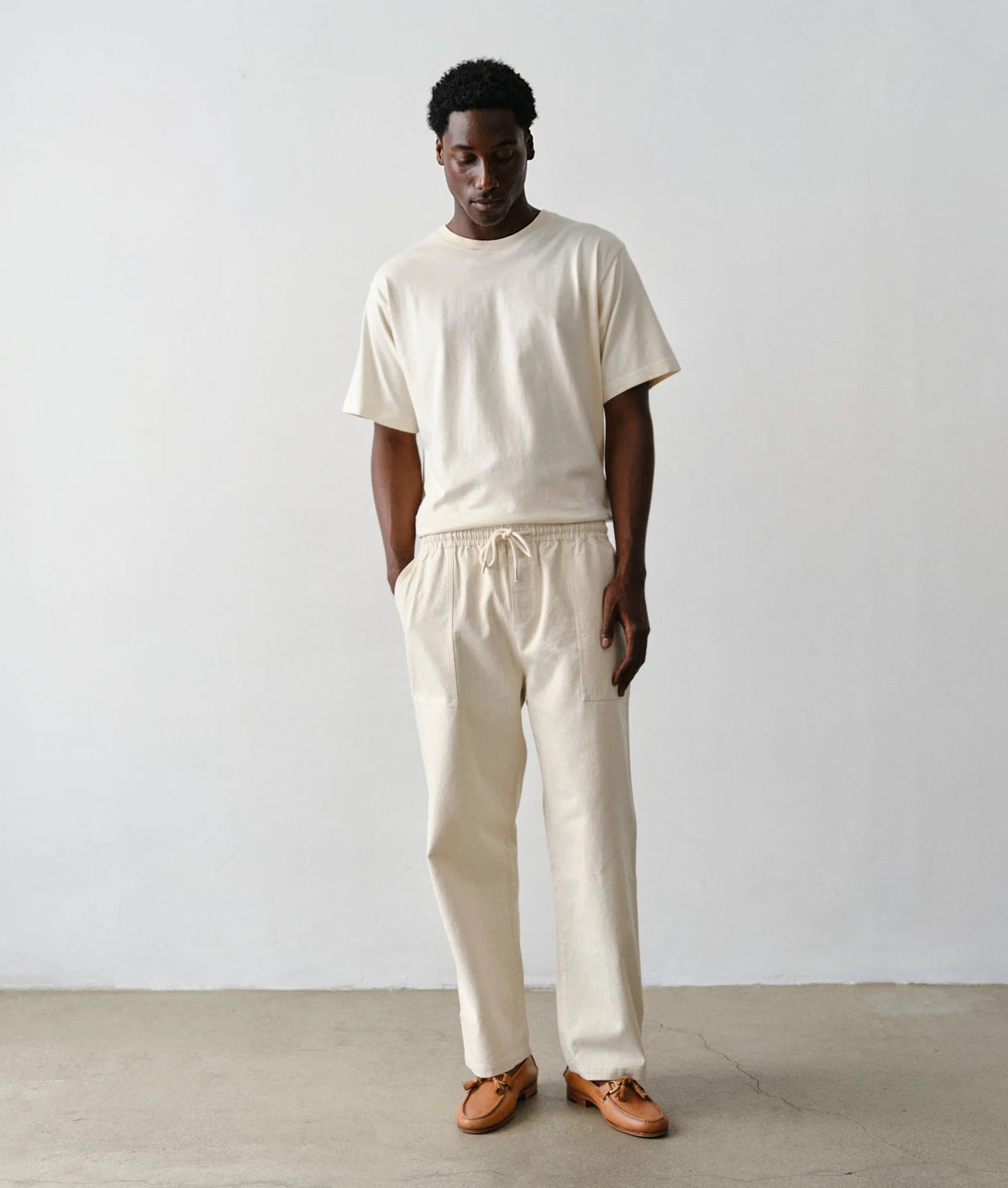 A man wearing IOAN undyed sleeve t-shirt and matching cream drawstring pants, paired with brown leather loafers, standing against a plain light gray wall.
