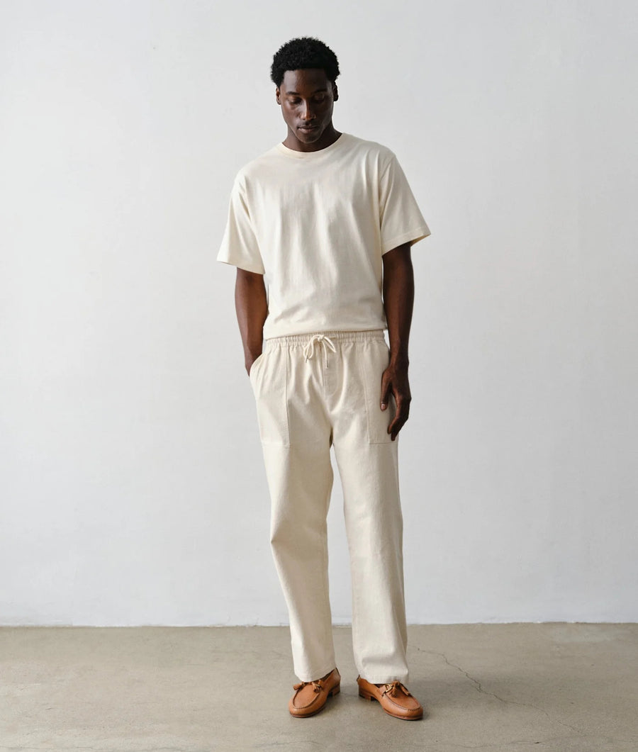 A man wearing IOAN undyed sleeve t-shirt and matching cream drawstring pants, paired with brown leather loafers, standing against a plain light gray wall.