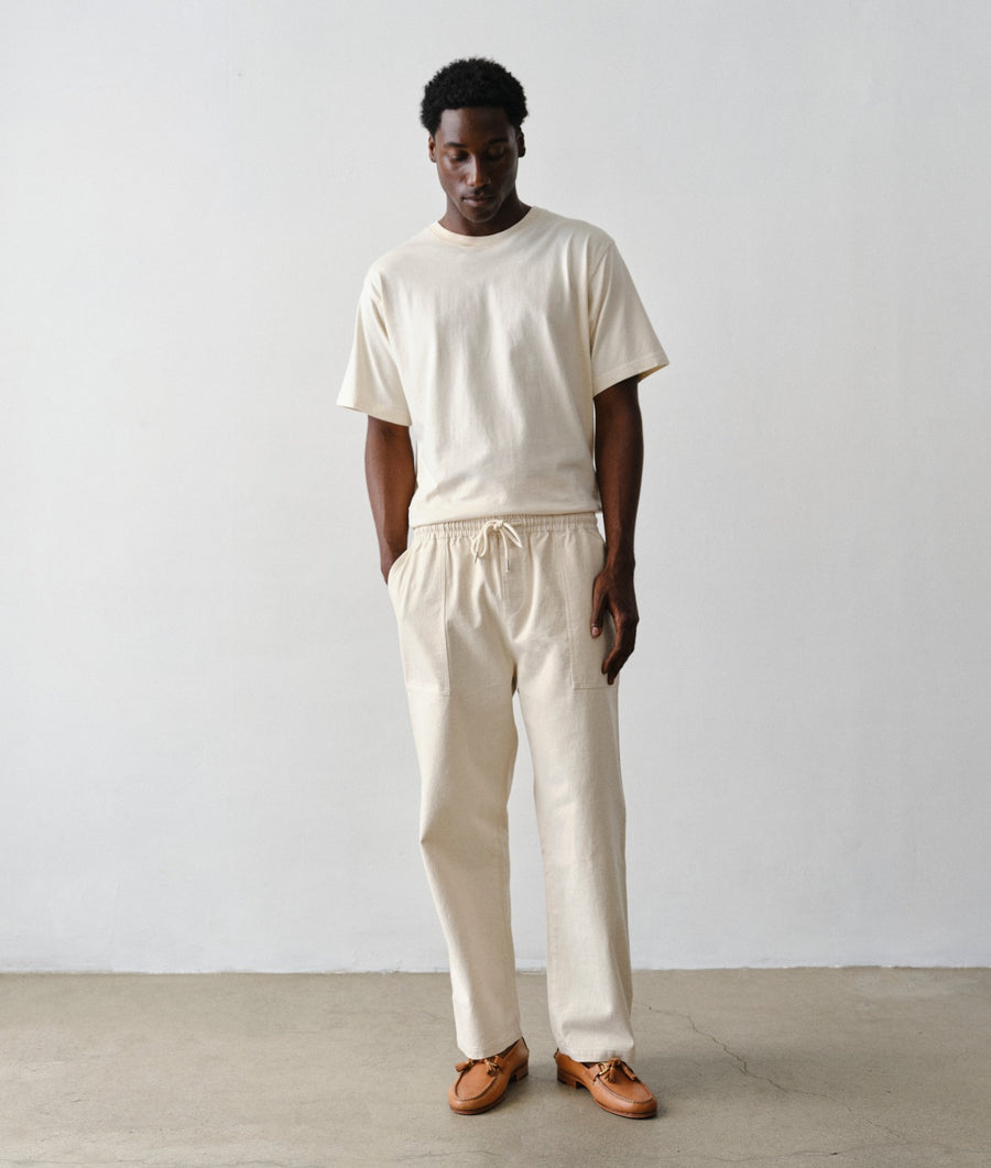 A man wearing an IOAN Clean Crewneck T-shirt with matching undyed Ango Pants, paired with brown leather loafers, standing against a plain light gray wall.
