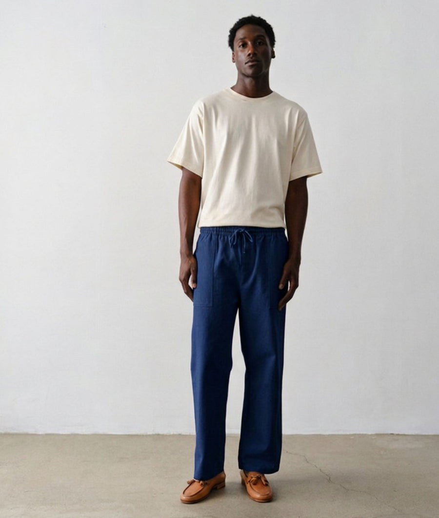 A man wearing IOAN Ango Pants in Indigo 12 and an IOAN Clean Crewneck T-shirt in Undyed stands against a plain light gray wall.