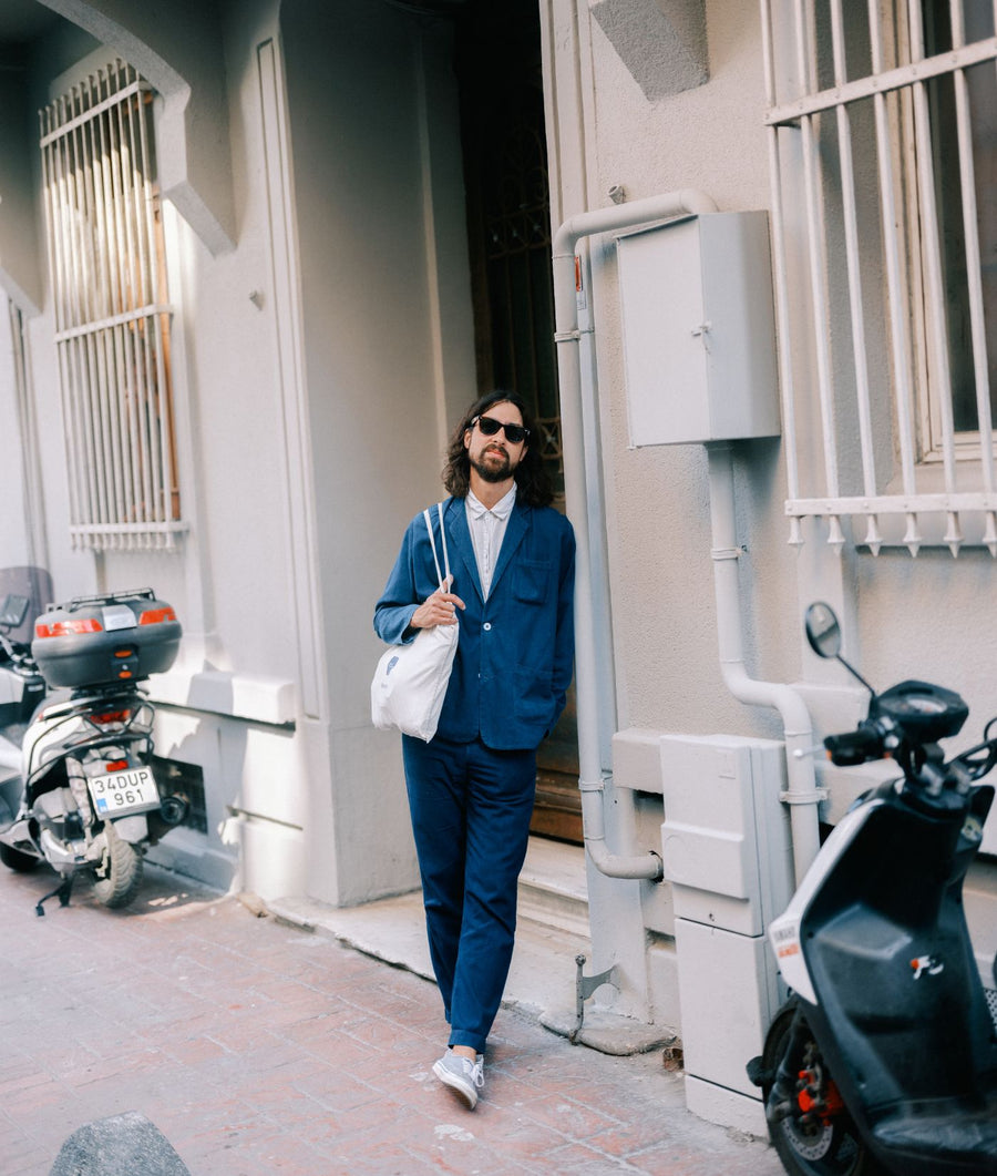 A man in a blue suit jacket, white shirt, and sunglasses, carrying a white bag, standing outside a building with a motorcycle parked nearby - Industry of All Nations