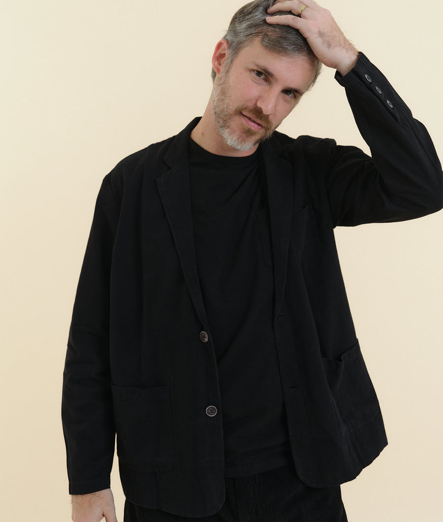 A man wearing a black wool blazer, with a contemplative expression - Industry of All Nations