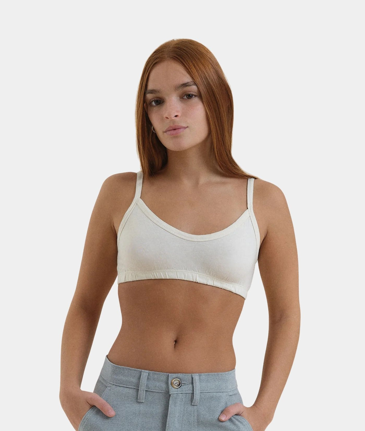 A woman with red hair wearing a cream-colored ribbed scoop-neck camisole crop top and light blue jeans, standing against a solid light gray background.