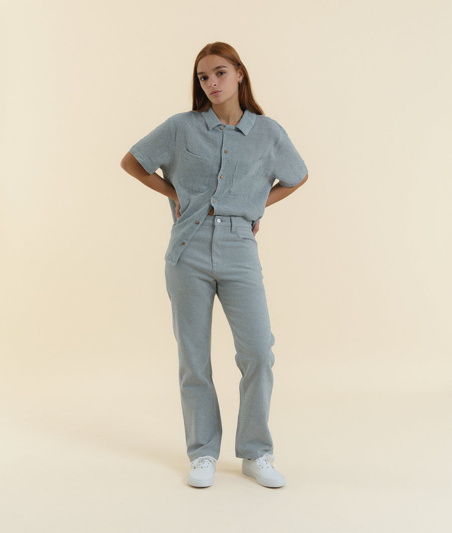 A young woman wearing a gray button-up shirt and matching pants, standing in a neutral-toned studio setting. - Industry of All Nations