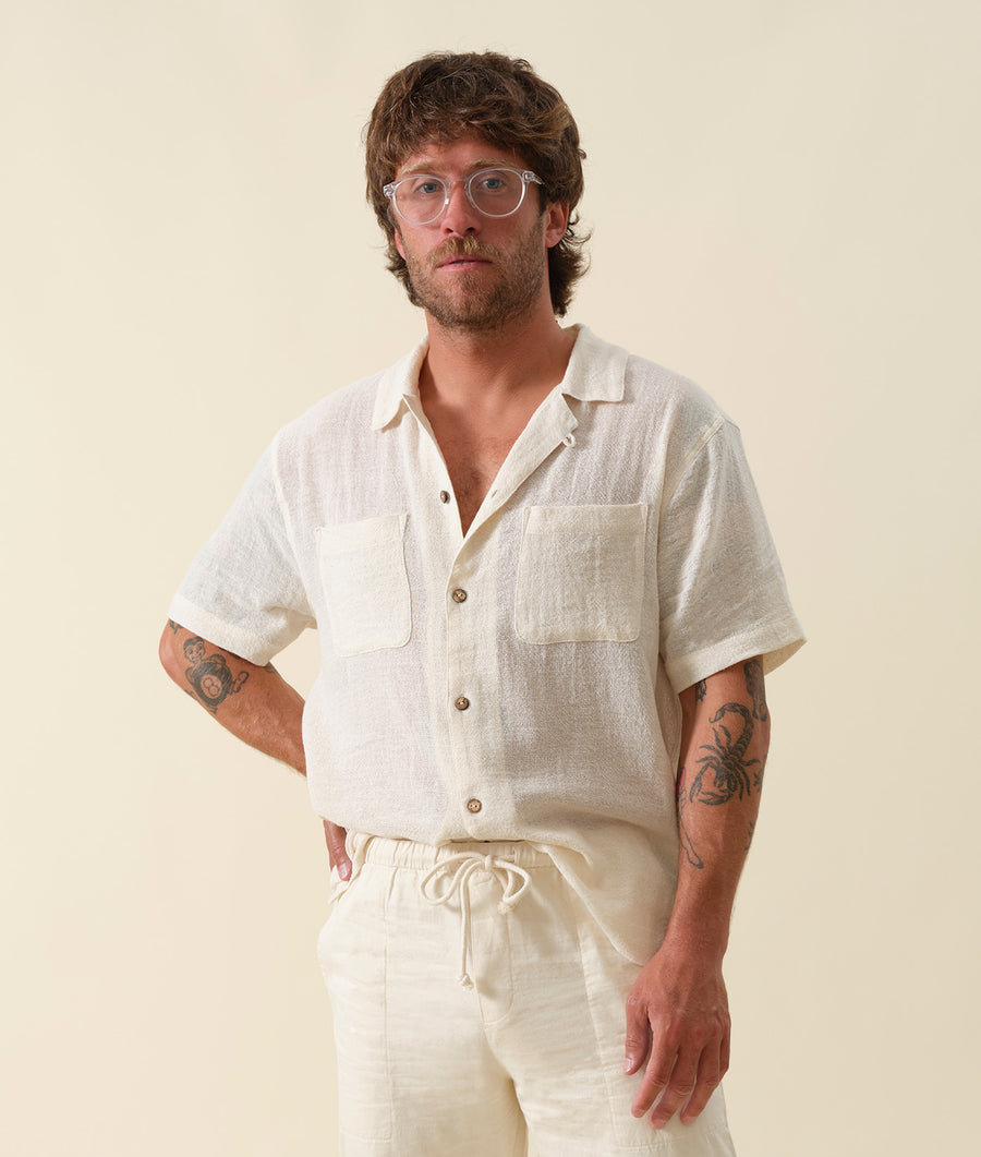 A man wearing a white linen button-down shirt with two front pockets, standing against a pale background - Industry of All Nations