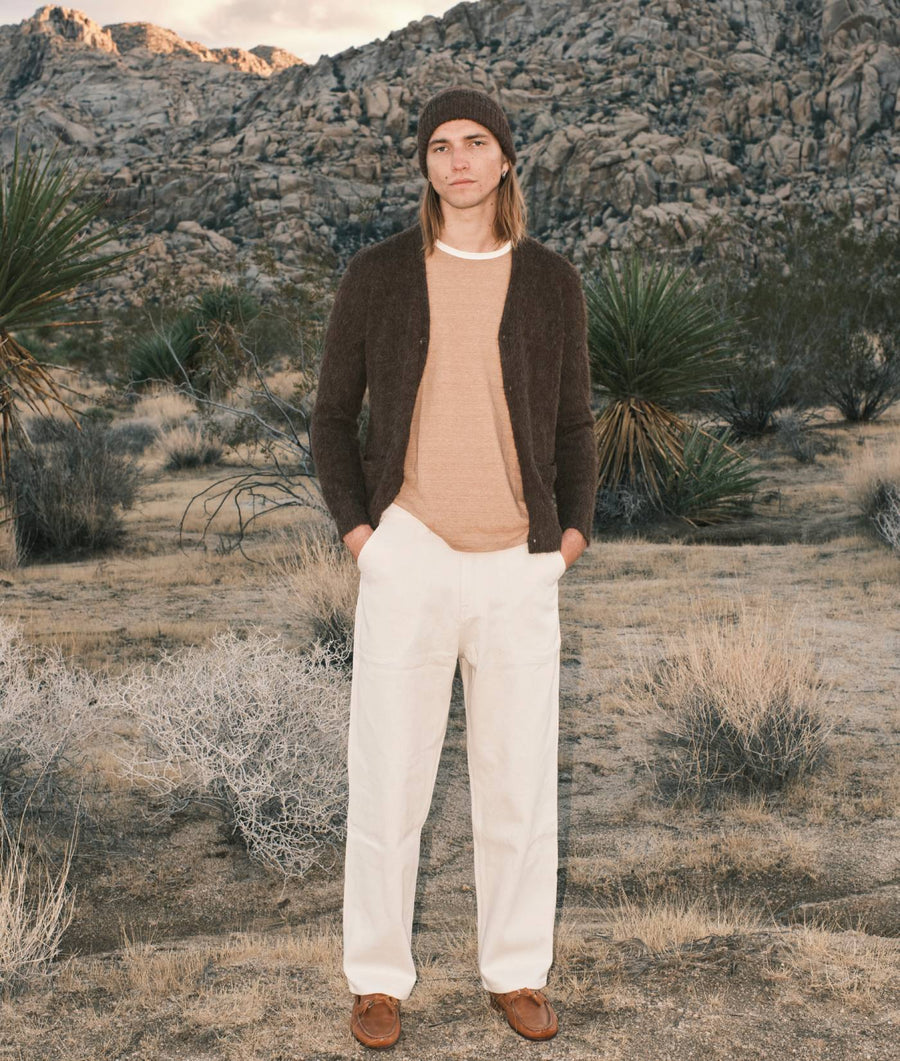 A person wearing a brown sweater, light colored shirt, and white pants, standing in a desert landscape - Industry of All Nations