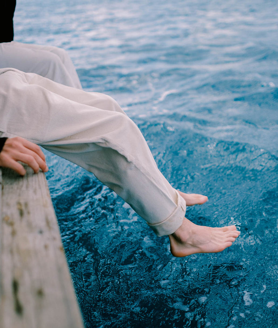 Feet dangling over the edge of a wooden dock, with the serene blue waters of the ocean visible in the background - Industry of All Nations