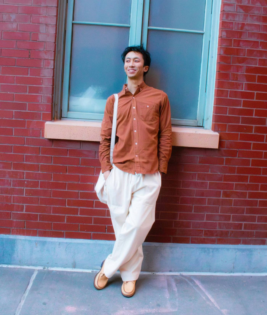 A smiling man wearing an orange button-up shirt and white pants, standing in front of a red brick wall - Industry of All Nations