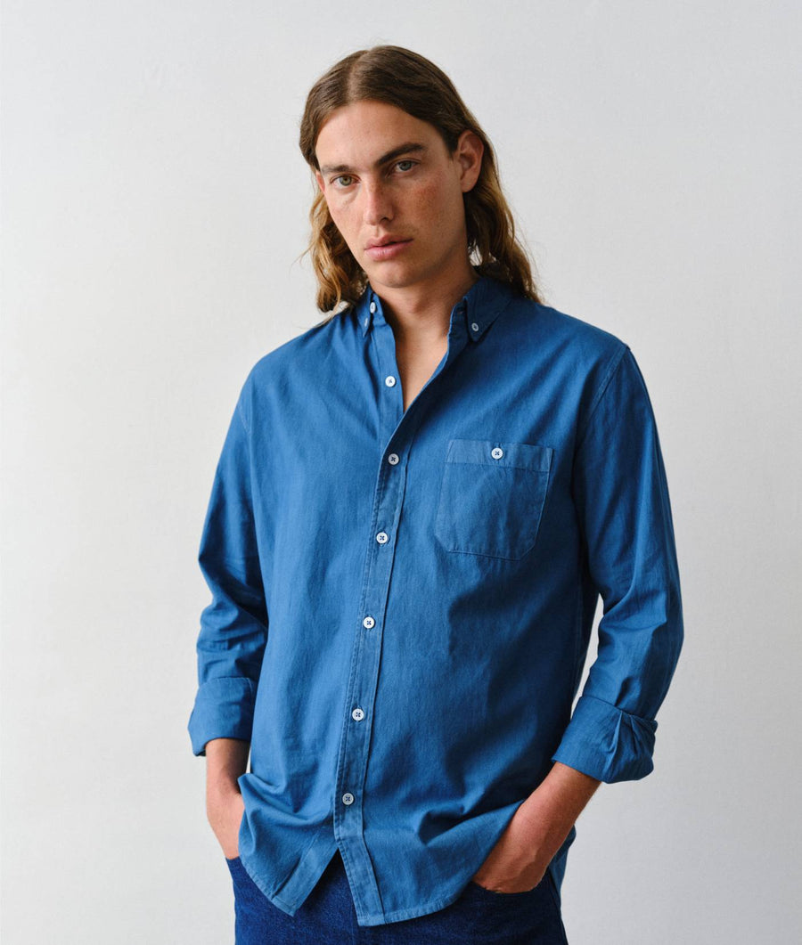 A man with long light brown hair wearing a blue long-sleeve button-down shirt with a chest pocket, posed against a light gray studio background.