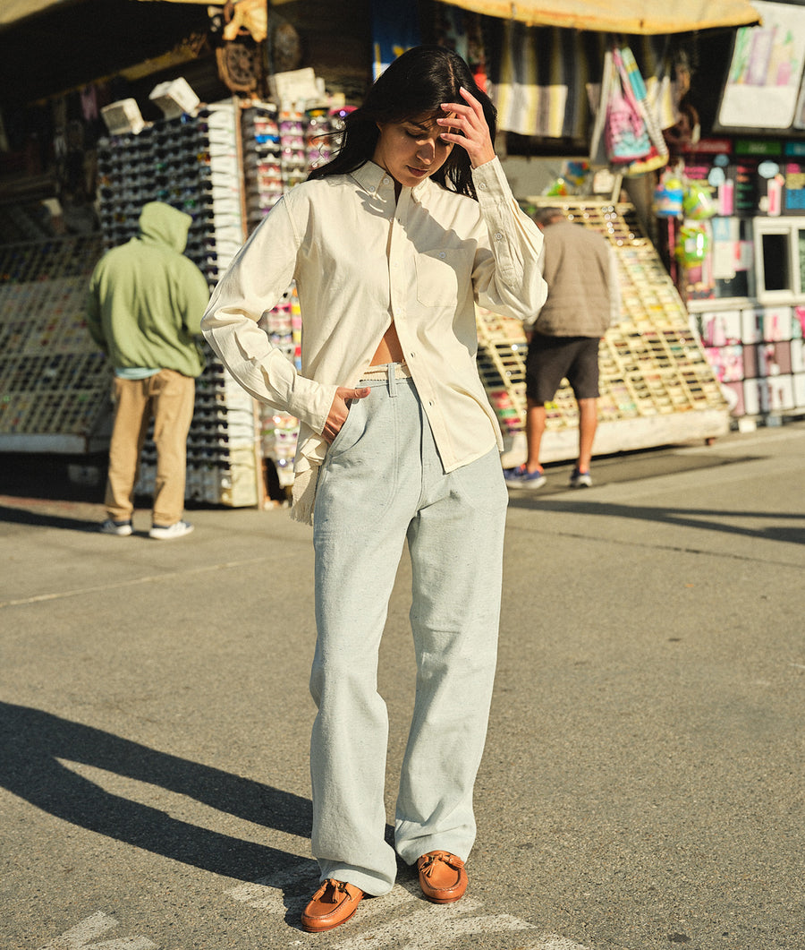 A woman wearing a white collared button-down shirt and gray wide-leg pants, standing in front of a colorful storefront - Industry of All Nations