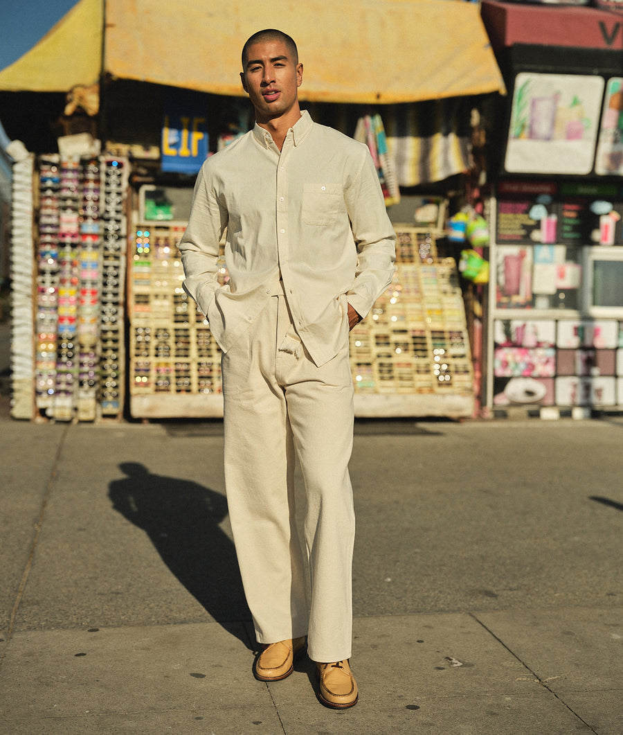 The image shows a male model wearing a light beige cotton jumpsuit with long sleeves and pockets, standing in front of a colorful retail store backdrop. - Industry of All Nations