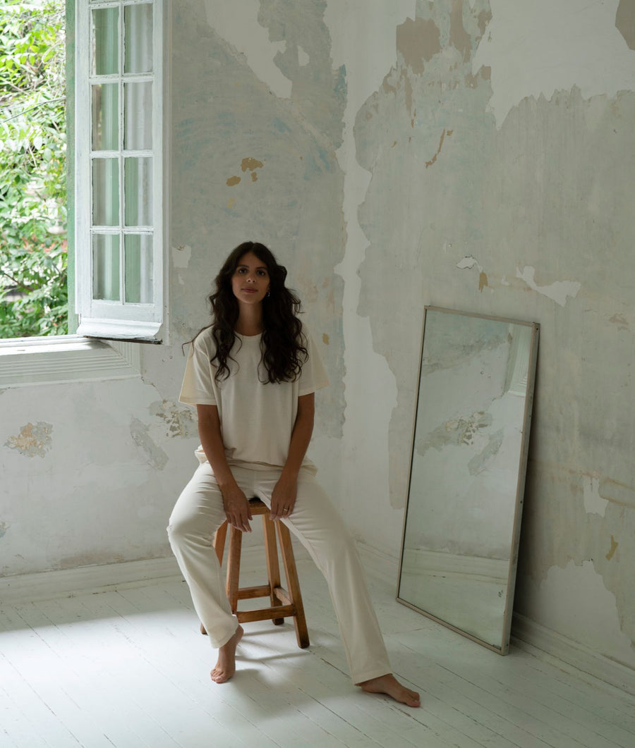 White t-shirt and pants worn by a person with long, dark hair sitting on a wooden stool in a rustic interior setting - Industry of All Nations