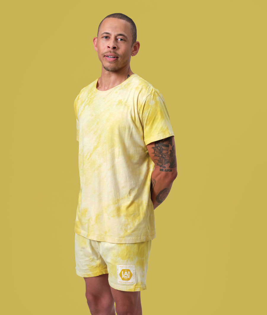 A young man wearing a yellow tie-dye t-shirt and shorts with a graphic logo on the left sleeve, standing in front of a yellow backdrop - Industry of All Nations