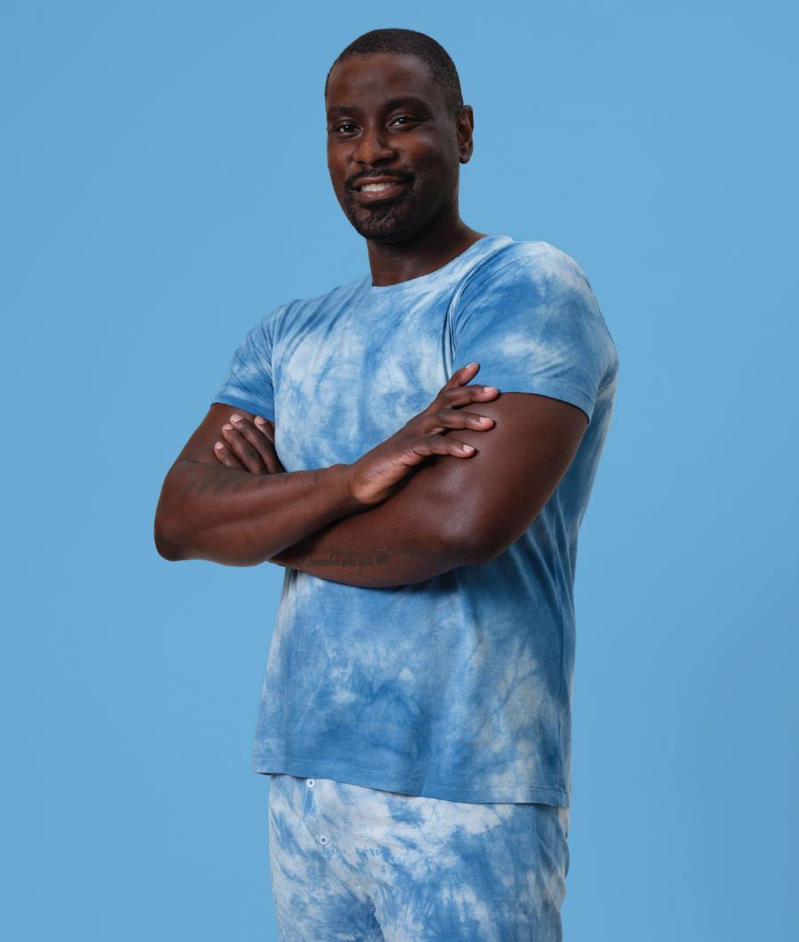 A young man in a blue tie-dye t-shirt and bottoms, standing with arms crossed against a blue background - Industry of All Nations