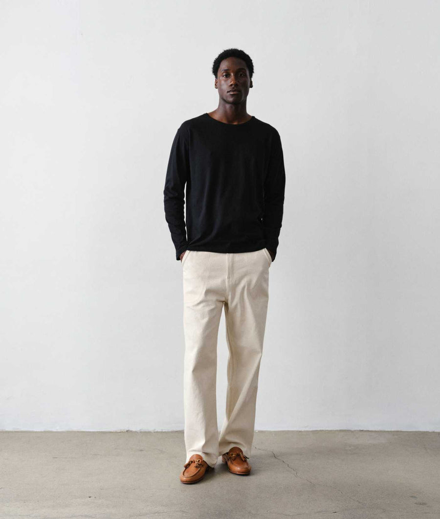 A man stands against a plain light gray wall wearing a black long-sleeve crewneck t-shirt, off-white trousers, and brown leather loafers.