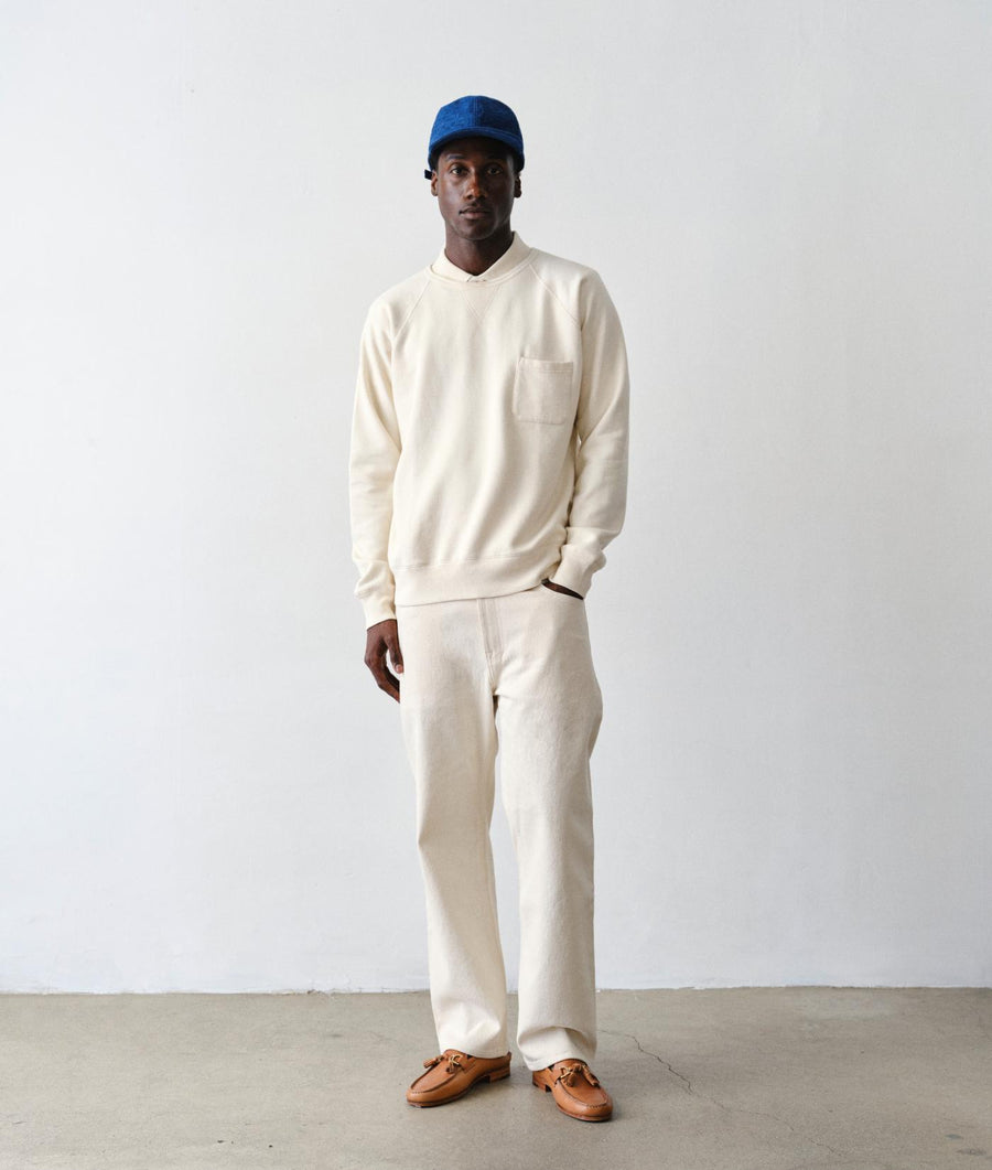 A man stands in a studio wearing a cream-colored knit sweater and matching wide-leg trousers, paired with a blue baseball cap and brown leather loafers against a plain white wall and concrete floor.