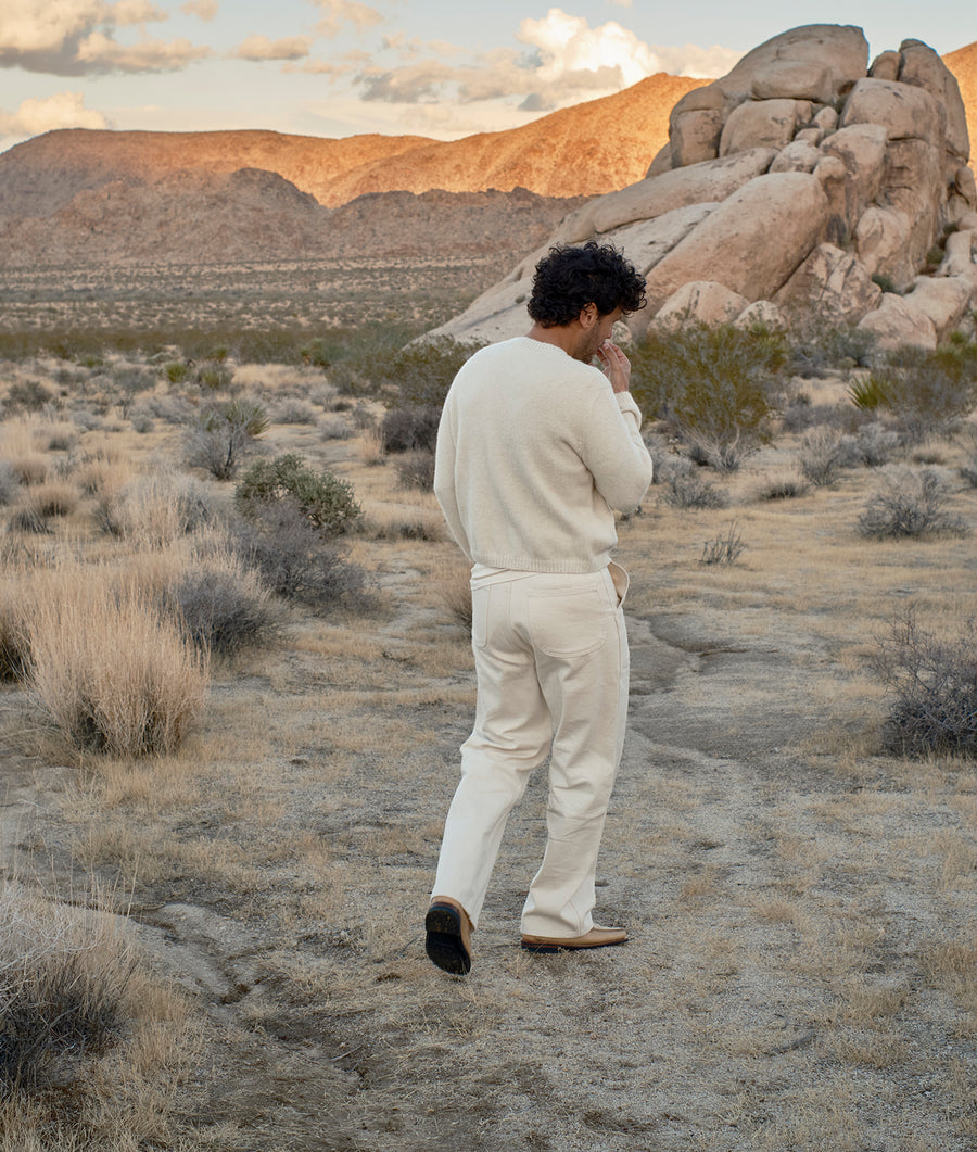A man wearing an off-white knitted sweater and pants, walking through a desert landscape - Industry of All Nations