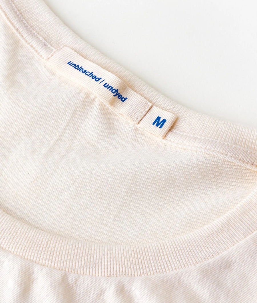 Closeup detail shot of a white cotton t-shirt with the text "Unbleached Undyed" in blue print, size tag "M" - Industry of All Nations