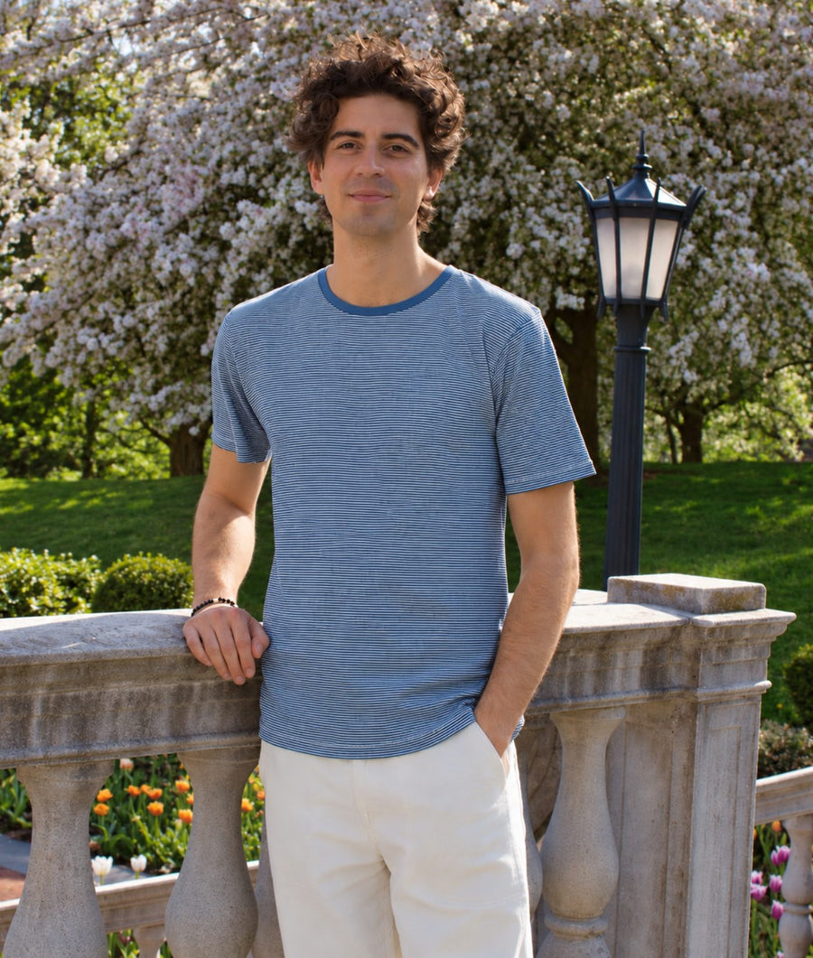 A man wearing a heathered slate blue t-shirt and off-white pants stands outdoors on a stone bridge with a blooming tree and a black lamp post in the background.