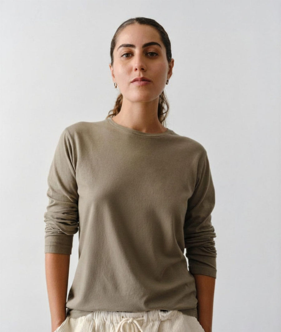 Woman stands against a plain light gray wall wearing an IOAN Iron 4 long-sleeve crewneck T-shirt.