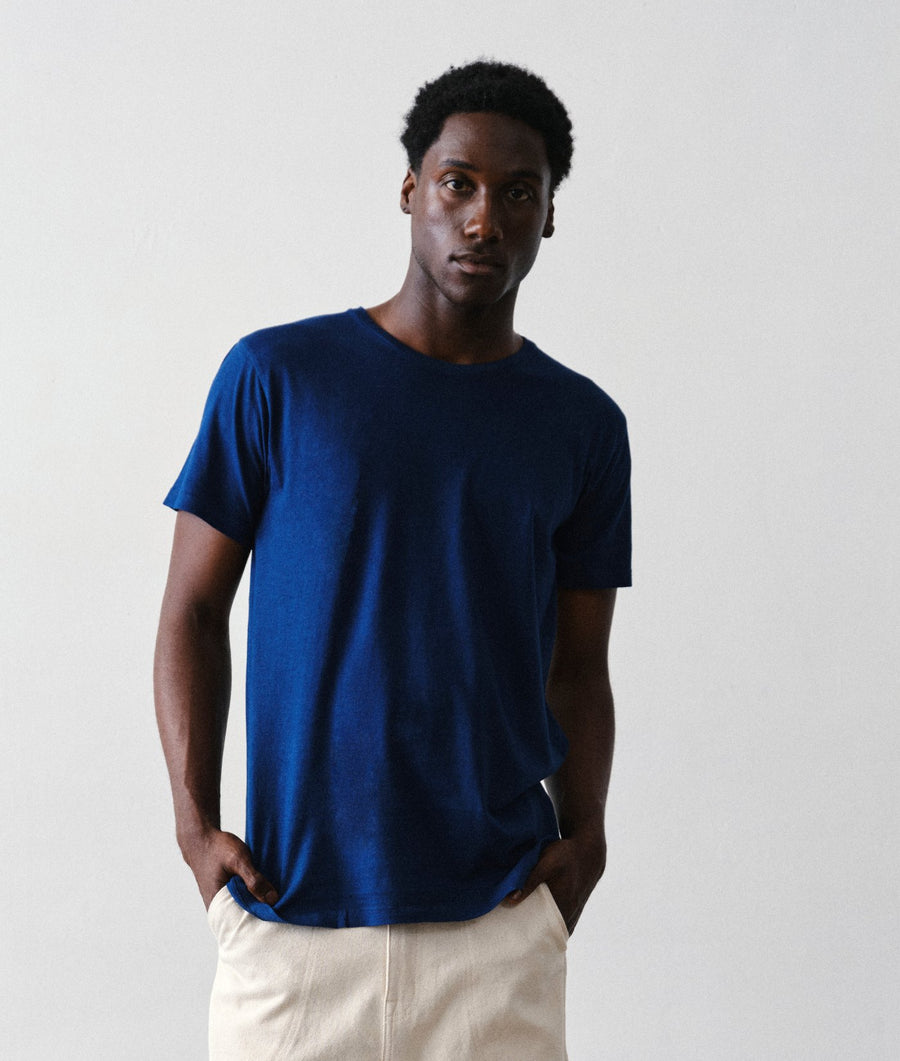 A man wearing the IOAN Indigo 12 Short-Sleeve Crewneck T-Shirt with undyed pants, standing against a light gray studio background.