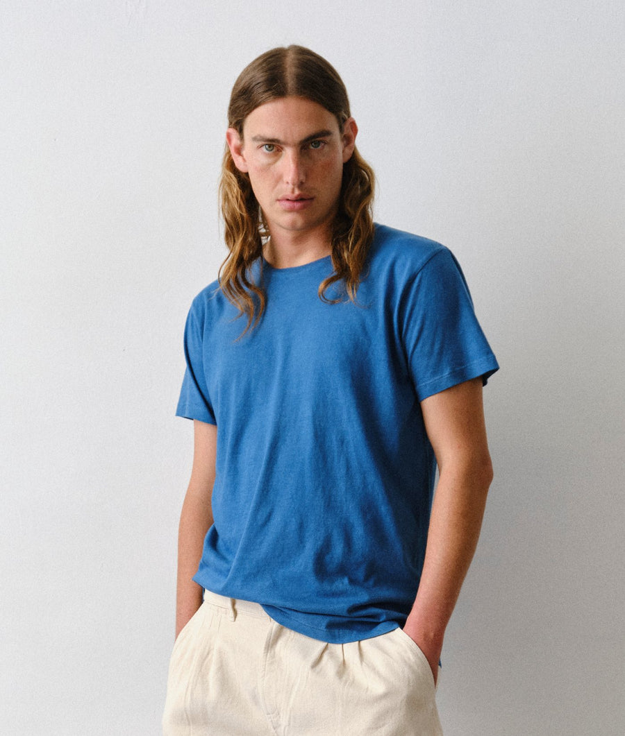 A man wearing the IOAN Indigo 6 Short-Sleeve Crewneck T-Shirt with undyed Gaucho pants, standing against a light gray studio background.