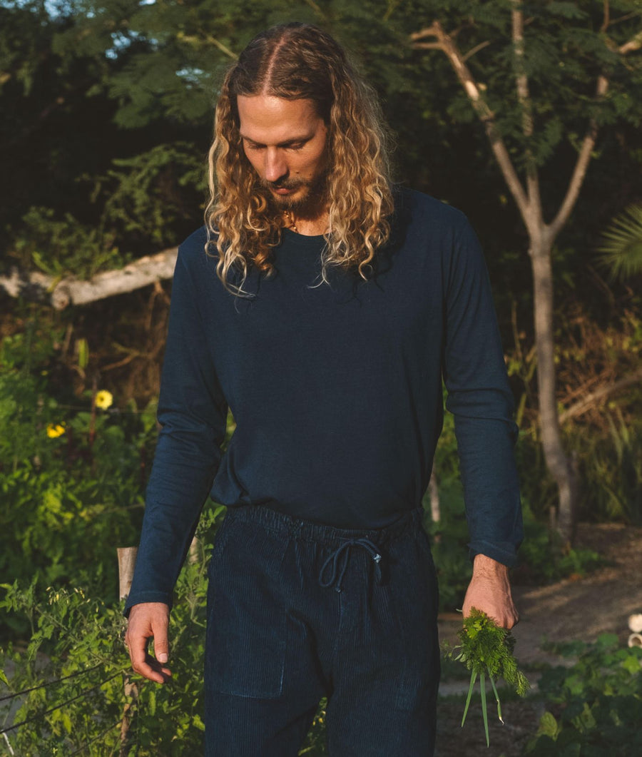 A man with long curly hair stands outdoors in a garden wearing an IOAN Clean Crewneck Long Sleeve T-shirt in Indigo 12 with matching corduroy pants, holding a small bunch of green herbs.
