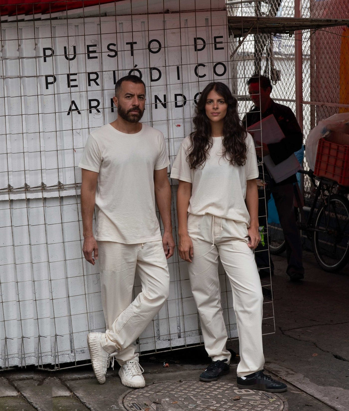 A man and a woman wearing matching IOAN Clean Crewneck T-shirts and Ponya Drawstring Pants in Undyed stand in front of a white tiled wall reading “PUESTO DE PERIÓDICO ARRIENDO.”